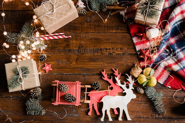 Christmas background with toy deer and sledge, giftboxes, pine cones, conifer, garlands and decorations on wooden table