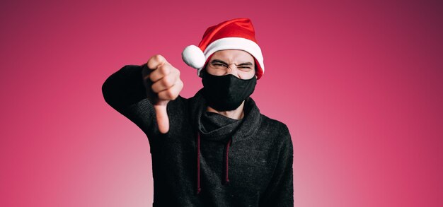 Christmas banner young man in black protective medical mask and\
red hat puts dislike with his hand