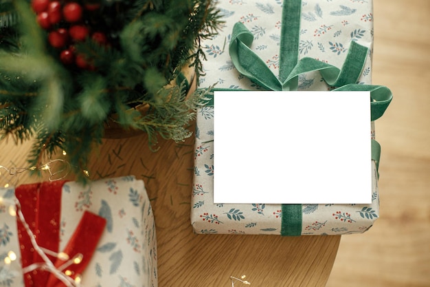 크리스마스 카드는 세련되게 포장된 크리스마스 선물과 나무 테이블에 축제 크리스마스 조명이 있는 전나무 가지에 빈 인사말 카드를 비웃습니다. 텍스트를 위한 공간 계절 인사말 엽서 템플릿