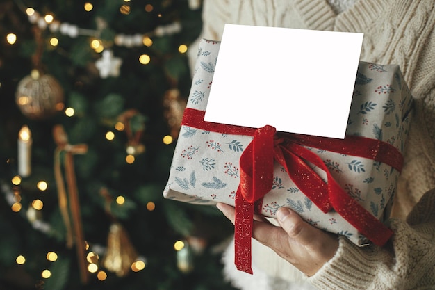 Modello di carta di natale mani che tengono una carta di auguri vuota e un regalo di natale avvolto in stile contro un albero di natale festivo con luci spazio per il testo modello di cartolina di auguri di stagione