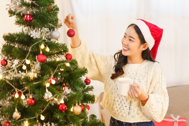 크리스마스 축하 개념 아시아 여성은 커피 컵을 들고 크리스마스 트리에 빨간 공을 장식합니다.