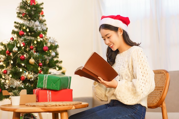 クリスマスのお祝いのコンセプト 若いアジアの女性はクリスマス ツリーと読書の近くの椅子に座っています。