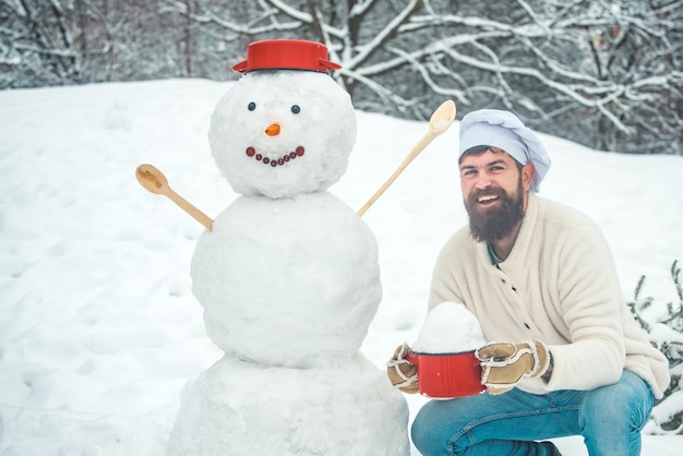 クリスマスシェフは雪だるまと雪の庭でハンサムな料理人の冬の肖像画を調理します