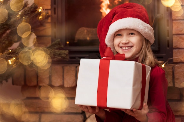 Christmas child girl in santa hat with big gift box or red\
present near christmas tree and fireplace