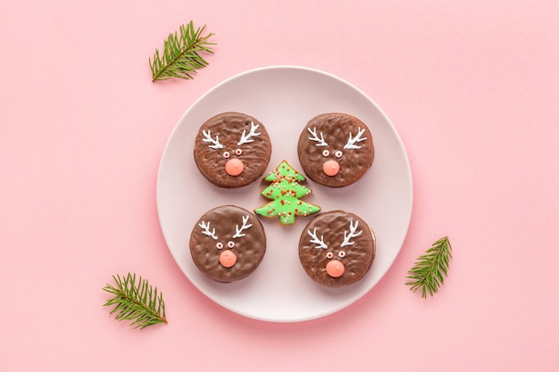 Christmas chocolate cookies decorated with funny deer faces around tree shaped gingerbread on dish standing on pink background.