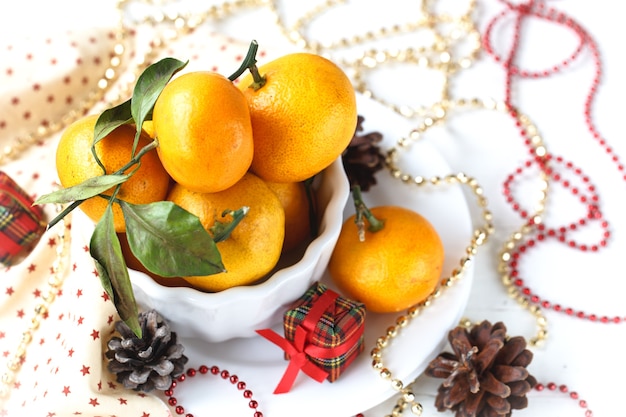 Новогодняя композиция из мандарина в белой миске, конусе, гирлянде и свечах на белом фоне