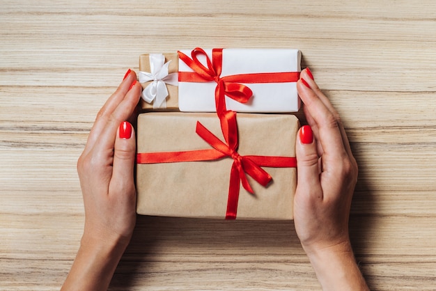 Photo christmas composition. top view, flat lay. female hands with red polished nails holding gift boxes wrapped in craft paper and decorated with satin ribbon on wooden table.