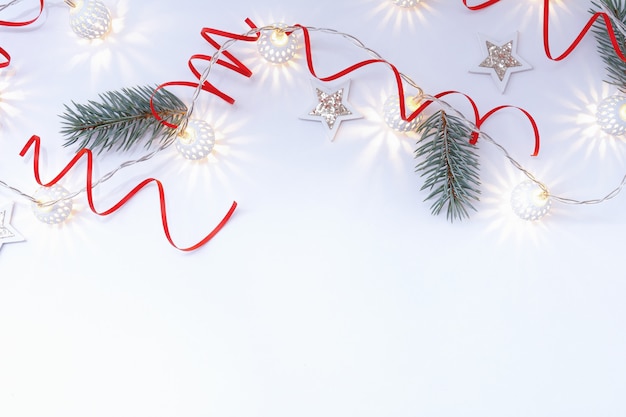 Christmas composition of white shiny stars, a garland of white luminous balls, red ribbons and fir branches on a white background.