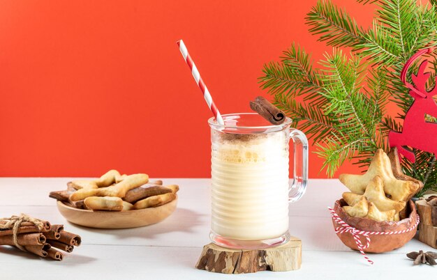 Christmas composition with eggnog in glass mug with ginger cookies on wooden table on red backdrop.