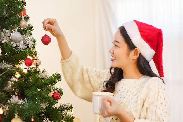 크리스마스 개념 커피 컵을 마시고 빨간 공을 들고 있는 아시아 여성이 크리스마스 트리를 장식합니다.