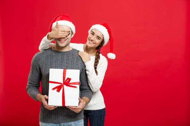 Christmas Concept - Jonge vrouw die de ogen van de man behandelt met de hand en een groot verrassingsgeschenk geeft. Geïsoleerd op rode muur.