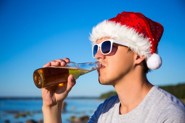 Christmas concept - portrait of young man in santa hat drinking beer on the beach