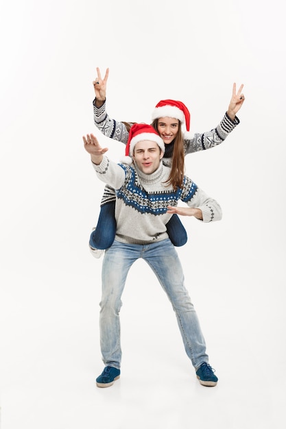 Концепция Рождества - молодая счастливая пара в свитерах, наслаждаясь контрейлерной поездкой, изолированной на белой серой стене.