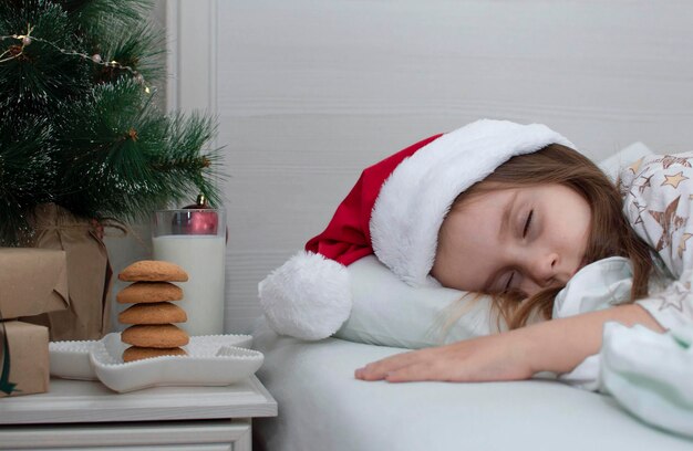 Christmas cookies and a glass of milk are a traditional treat. A charming little girl fell asleep on the bed near the bedside table with a treat for Santa Claus. The concept of the New Year. Relatives