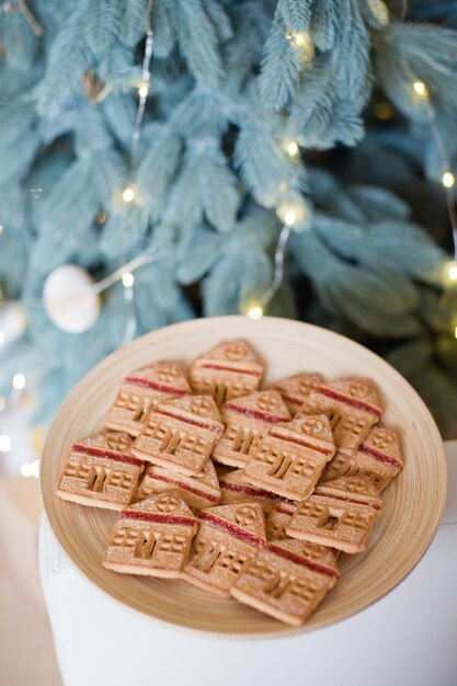 Christmas cookies house on a wooden plate near Christmas tree with lights and decorations Christmas sweets homemade bakery