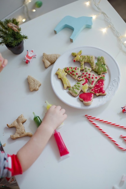 カラフルな氷の釉薬と白いプレート上のクリスマスクッキー子供の手はクッキーを取りますクリスマスクッキーの背景チューブで焼いたクッキー釉薬キャンディー杖とライト