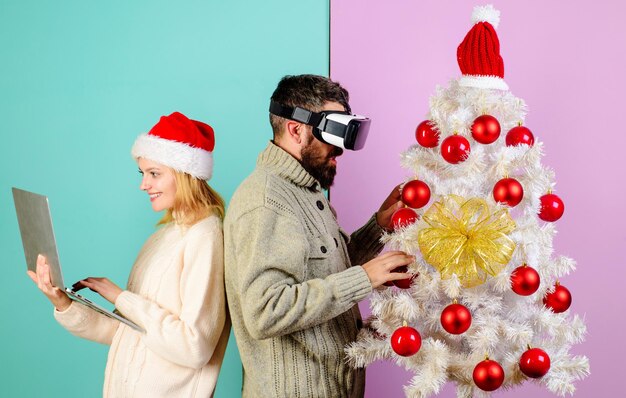 크리스마스 부부는 VR 안경을 입은 수염이 있는 남자와 크리스마스 근처에 노트북을 가진 산타 모자를 입은 웃는 여자