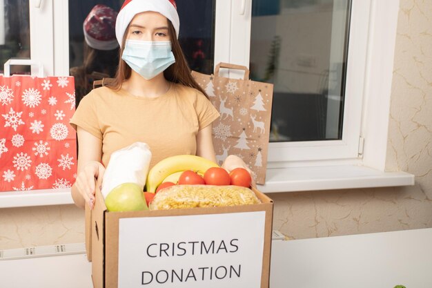 Foto donazione covid di natale una giovane volontaria che indossa una maschera medica in un centro di beneficenza raccoglie cibo in una scatola per le vittime della pandemia di coronavirus