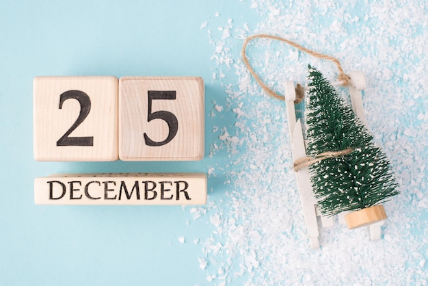 Photo christmas day on calendar concept. top above overhead close up photo of wooden cubes with date and toy miniature tree on white wooden sledges sleigh