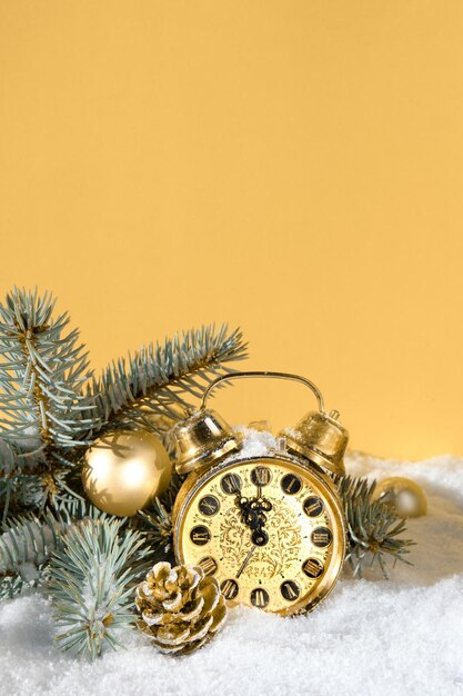 Christmas decor, gold alarm clock on golden yellow background. Sunshine, snow and fir twigs.