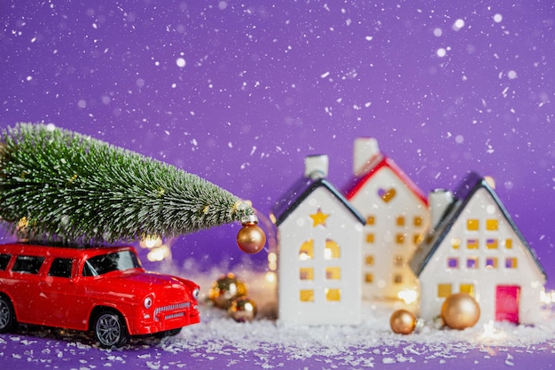 クリスマスの装飾-雪の上の赤いレトロな車は、屋根にギフトボックスが付いたボケのクリスマスツリーにフェアリーライトが付いた家を通り過ぎます。紫の背景のおもちゃ。新年のグリーティングカード。居心地の良い家