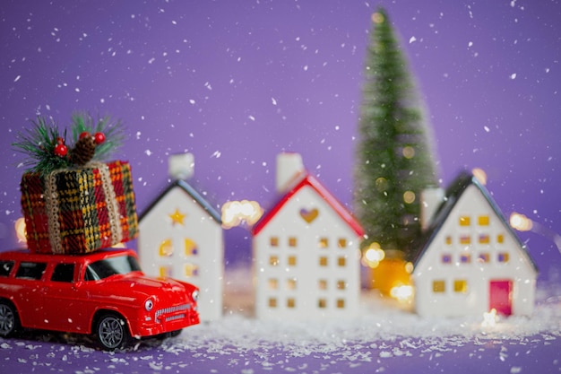 クリスマスの装飾-雪の上の赤いレトロな車は、屋根にギフトボックスが付いたボケのクリスマスツリーにフェアリーライトが付いた家を通り過ぎます。紫の背景のおもちゃ。新年のグリーティングカード。居心地の良い家