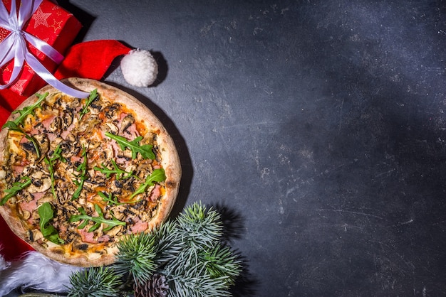 ペパロニとキノコのピザでクリスマスに飾られた背景、配達とレストランのクリスマスランチとパーティーメニュー、サンタはクリスマスにピザを配達します、黒いコンクリートの背景上面図コピースペース