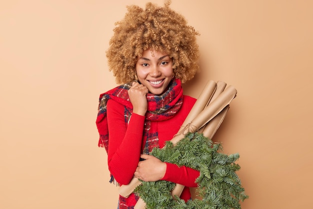 Decorazione e composizione natalizia. felice dai capelli ricci giovane donna indossa sciarpa rossa del ponticello intorno al collo porta la carta ghirlanda di abete verde arrotolata isolata su sfondo beige. atmosfera festosa