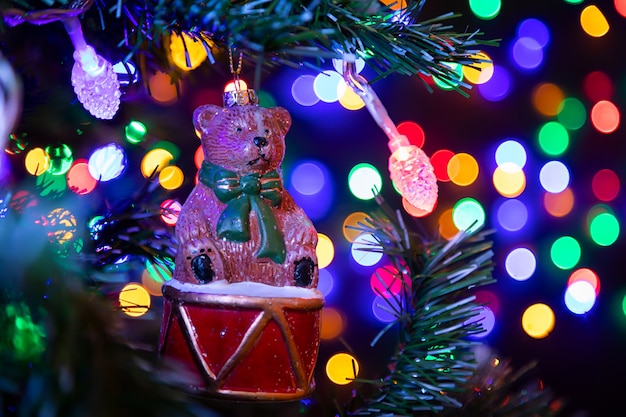 Christmas decoration in the form of a bear on a drum hanging on a Christmas tree in the background many garlands in different colors glow. 