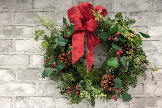 Corona della decorazione di natale con le bacche rosse dell'agrifoglio, i coni di abete e l'arco rosso del nastro sul muro di mattoni