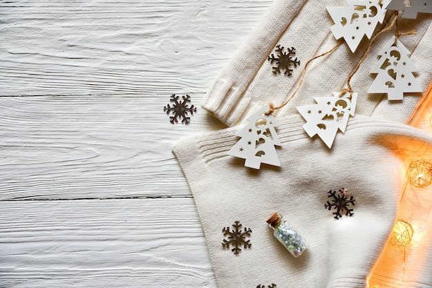 白い木製の背景のクリスマスの装飾。白い毛糸の帽子とスカーフ、レースに白い木のモミの木、銀の雪片、装飾的な照明、輝きのある装飾的なボトル。