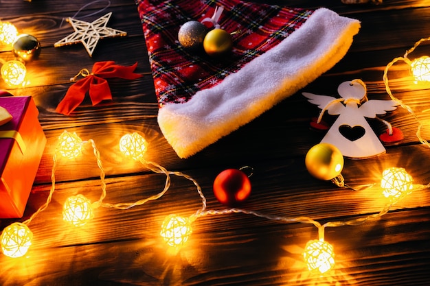 Christmas decorations on the wooden table. Lights, baubles and Santa hat. Evening romantic mood. Cozy atmosphere. Holidays flat lay. Festive concept. New year 2021.