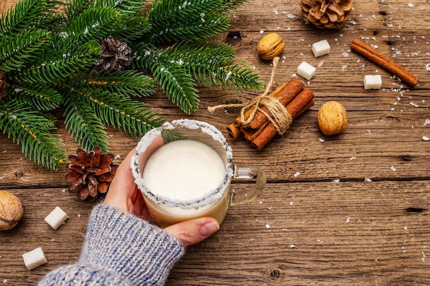 Christmas eggnog liqueur or cola de mono cocktail. Classical winter drink in glass mug, xmas decorations. Woman's hand in jersey.