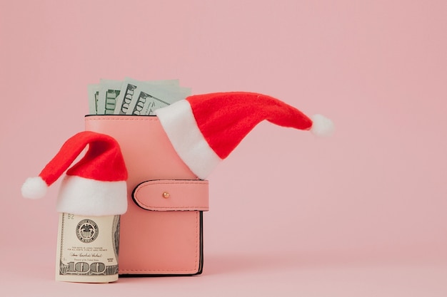 Christmas expenses. Pink leather purse with santa claus cap, gift, fir tree and dollars banknotes on pink . Christmas shopping. Holiday Sale