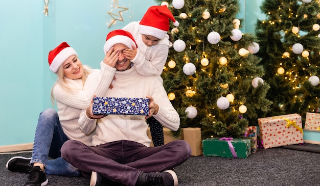 クリスマス。家族。幸せ。カメラを見て、家の床に笑みを浮かべてサンタの帽子をかぶったお父さん、お母さんと娘