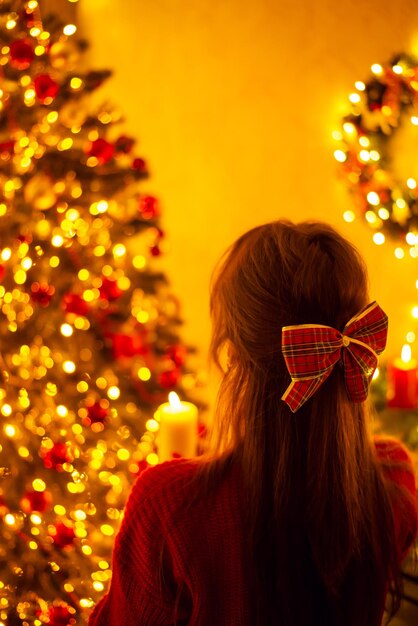 Christmas festive decorations girl in decorated room for\
celebrating new year with burning candle back shot of a dreamy\
beautiful girl admiring beauty of christmas lights in decorated\
room at home