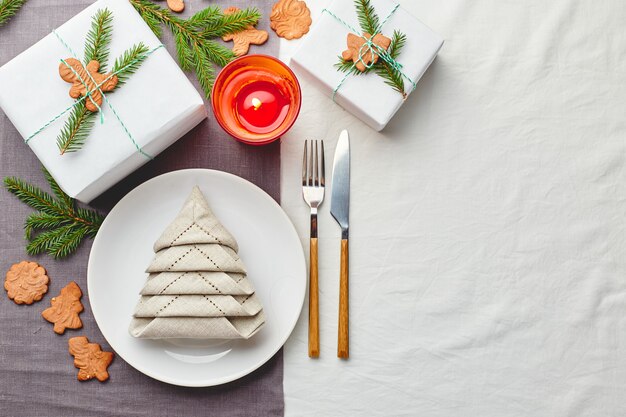 Christmas festive table setting, napkin in the form of Christmas tree on a plate on white tablecloth with gifts and decorations with fir sprigs and gingerbread cookies