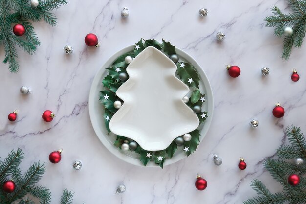 Christmas flat lay on marble table, copy-space on ceramic plate. Xmas background in green, white and red. Natural fir twigs, holly leaves and glass trinkets.