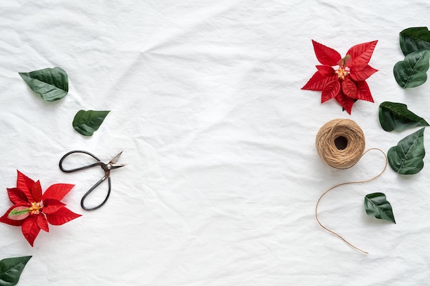 Christmas flat lay with copy-space. Red poinsettia, Xmas top view on white textile background, text space. Hemp cord, scissors, red and dark green poinsettia leaves .