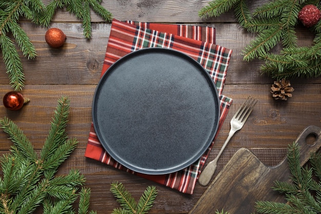 Christmas food border with black plate evegreen branches and red decor on wooden board Space for text Top view