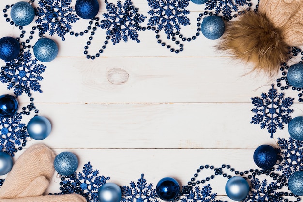 Christmas frame of blue balls, beads, stars and warm knitted hats and mittens on a white wooden table, top view, copy space. Winter, new year, holiday and decor concept