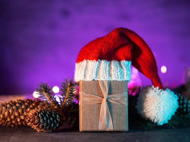 Новогодняя подарочная коробка в вязаной шапке Санта-Клауса на неоновом фоне. Ветка елки с шишками.