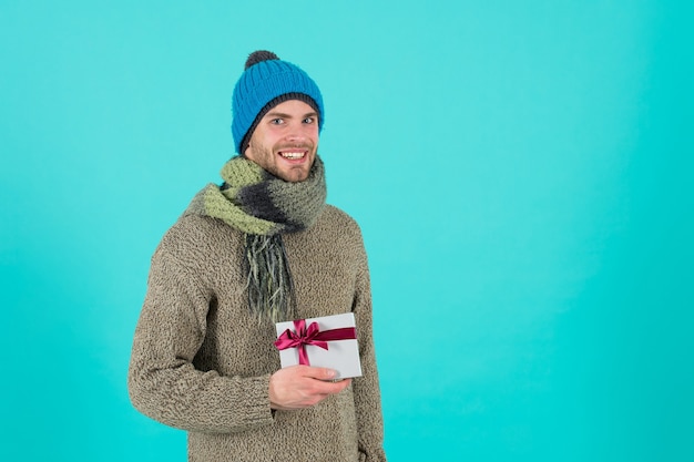 Christmas gift. Guy wear winter hat. Merry christmas and happy new year. Winter vacation. Man in hat face bristle. Positive emotions. Handsome man celebrate winter holidays blue background.
