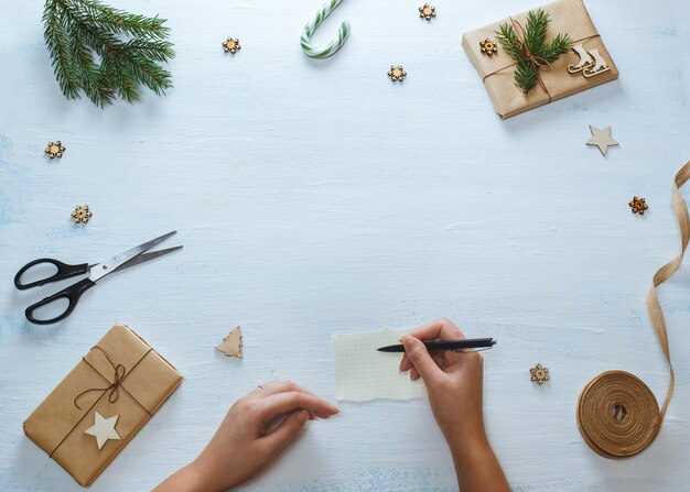 Christmas gift packaging composition. Female hands with pen, Christmas gifts, candy, fir branches, scissors, ribbon, on blue wooden background. Flat lay, top view