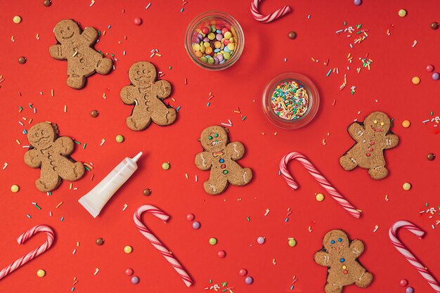 Christmas gingerbread cookie preparation process with gingerbread man, icing, sugar sprinkles and candy canes on a red table background. New year wallpaper flat lay.