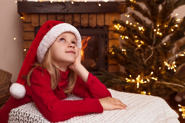 Christmas Girl Child Waiting for Santa and Dreaming on Christmas Eve near Tree and Fireplace. Space