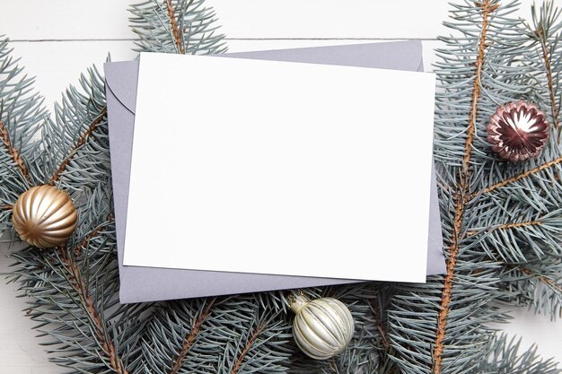Макет рождественской открытки с конвертом, зелеными еловыми ветками и шарами на белом деревянном фоне, вид сверху, плоская планировка, макет зимней праздничной открытки с украшениями