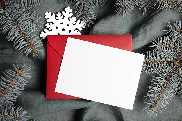 Christmas greeting card mockup with fir tree branches red envelope and white snowflake on green background top view