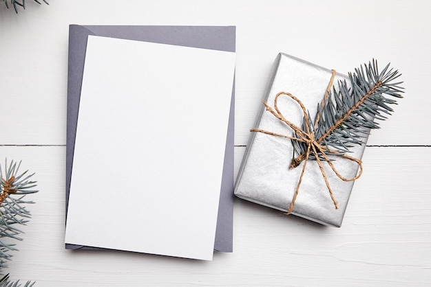 Макет рождественской открытки с серым конвертом серебристого цвета подарочной коробкой и зеленой еловой веткой на белом деревянном фоне вид сверху плоская планировка Пустая зимняя праздничная открытка