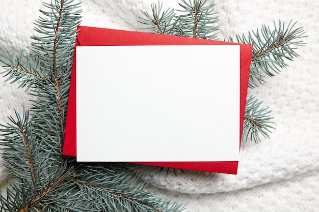 Christmas greeting card mockup with red envelope and fir tree branches on white knitted clothes background top view flat lay